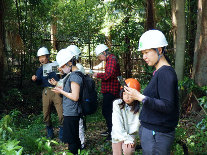 樹木が茂る森の中で、白いヘルメットを被った参加者たちがスマートフォンや資料を確認している様子。