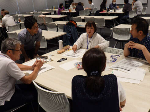明るい会議室で、テーブルを囲んだ5人の男女がグループディスカッションやワークショップを行っている様子