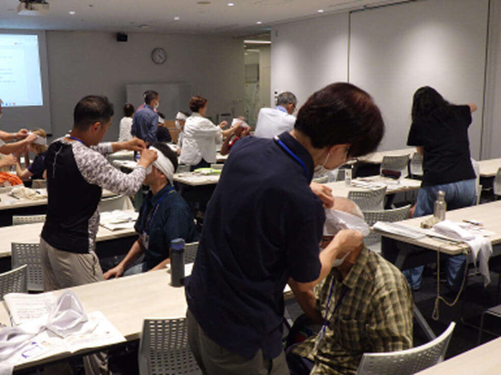 室内で行われている応急救命講習で、頭部への包帯の巻き方を実習している受講生たちの様子