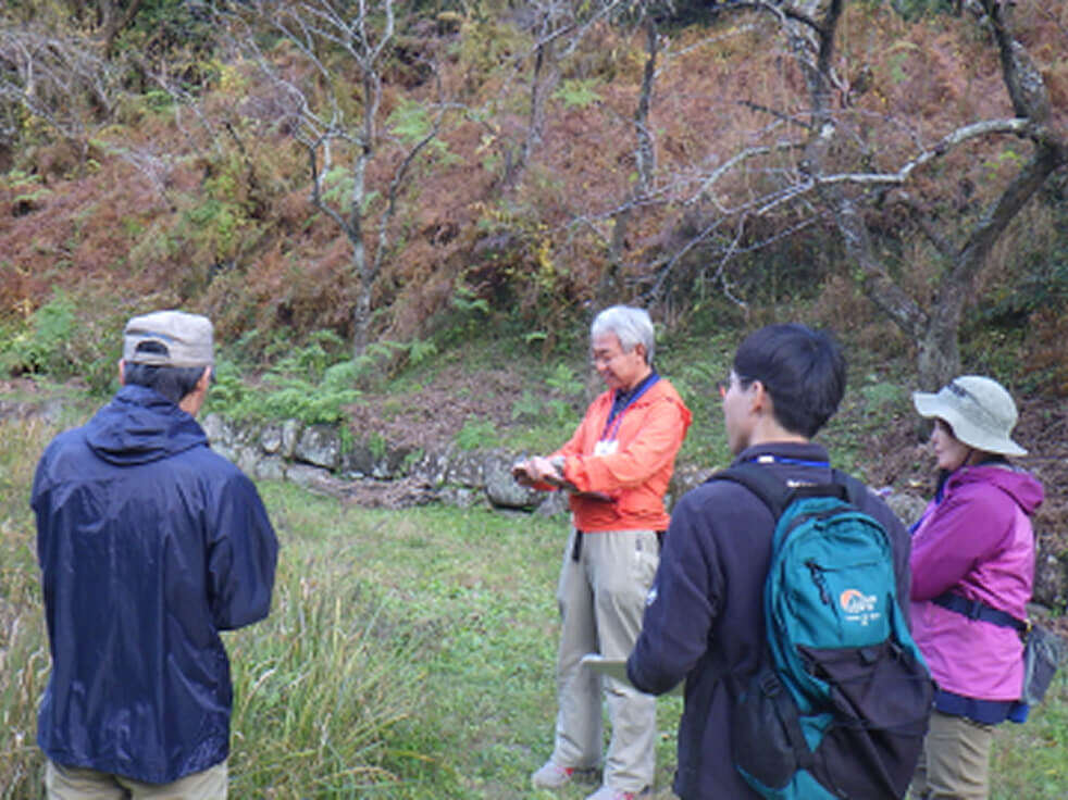 紅葉した木々やシダが生える斜面を背景に、オレンジ色のジャケットを着た講師が、3人の参加者に向けて自然観察の解説や指導を行っている様子