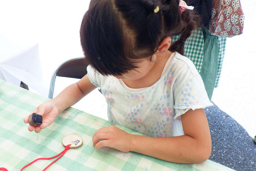 チェック柄のテーブルクロスが敷かれた机で、女の子が木材（輪切り）を使ったペンダント作りの工作をしている様子