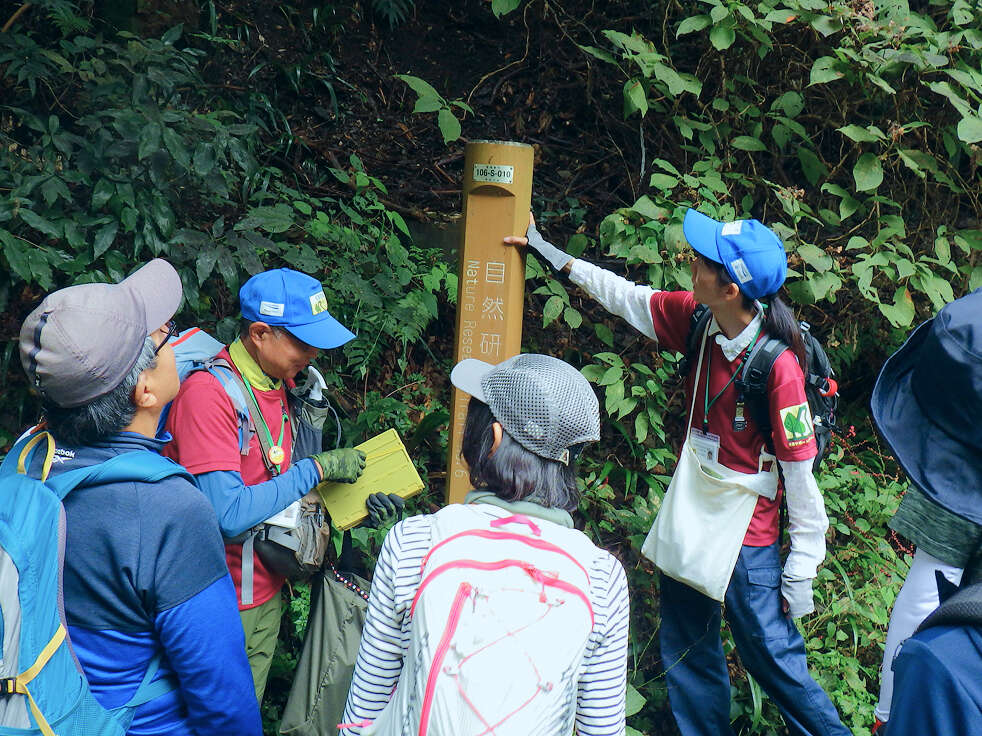 青い帽子を被ったスタッフが、森の中で登山客に対し、茶色の標識柱を指差しながら解説している様子。