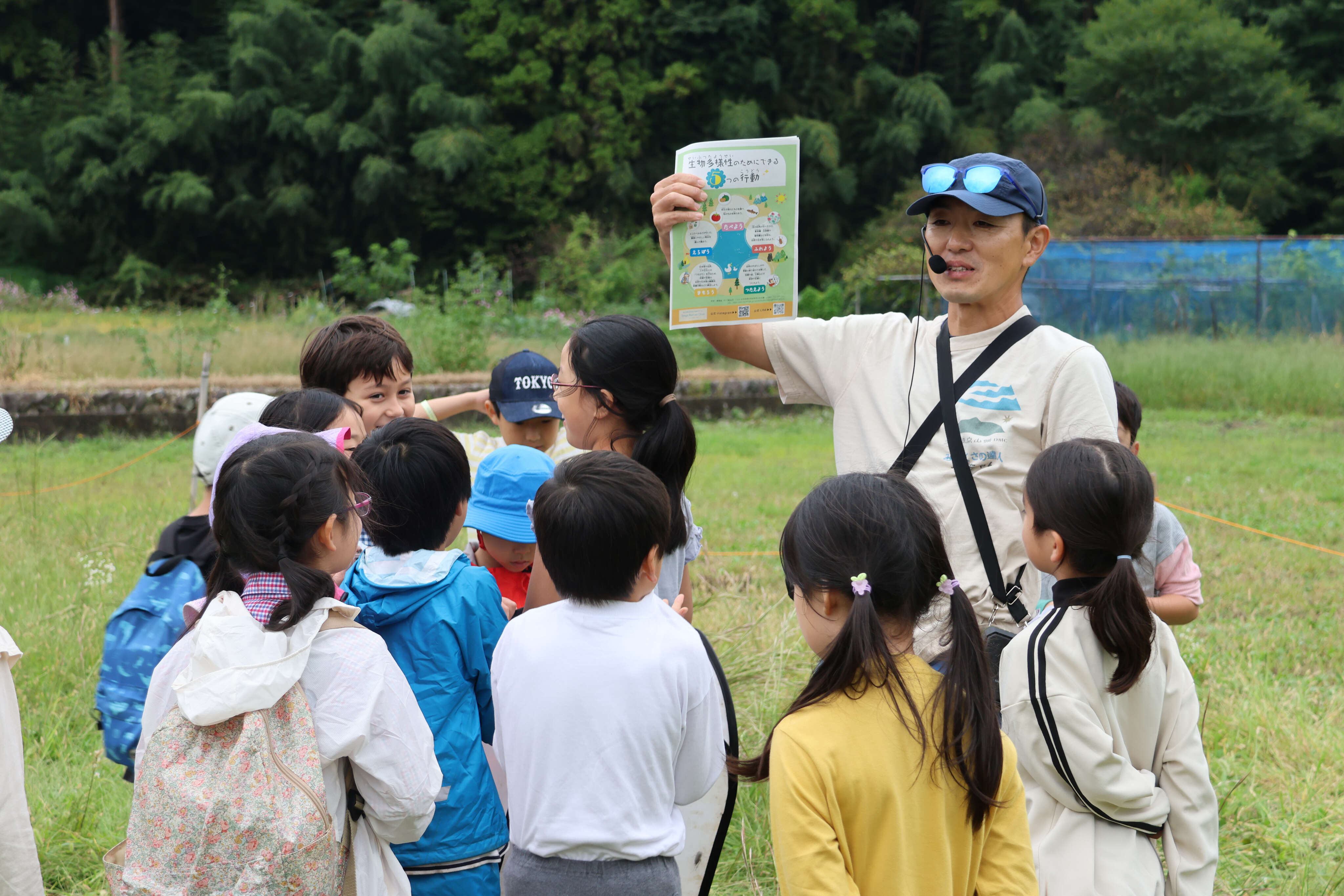 緑豊かな野外フィールドで、指導員の男性が「生物多様性のためにできる5つの行動」のチラシを掲げ、集まった子供たちに解説している様子
