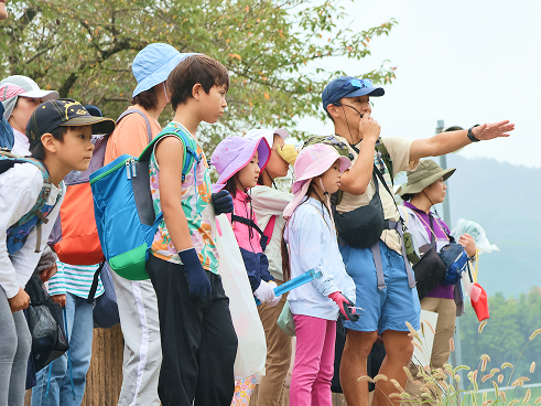 野外フィールドで、指導員の男性が指差す方向を、リュックを背負った子供たちが真剣な表情で見つめている様子