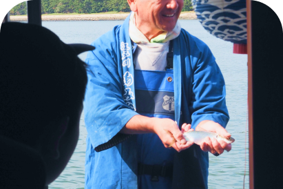 船の上で、青い法被を着た男性が、手にした小魚を参加者に見せている様子