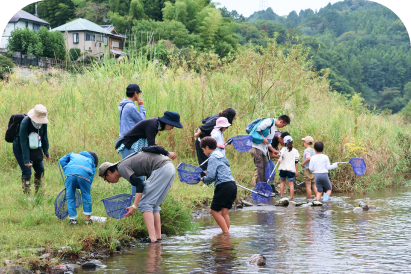 川岸で網を持って水辺の生き物を探す親子や参加者たち