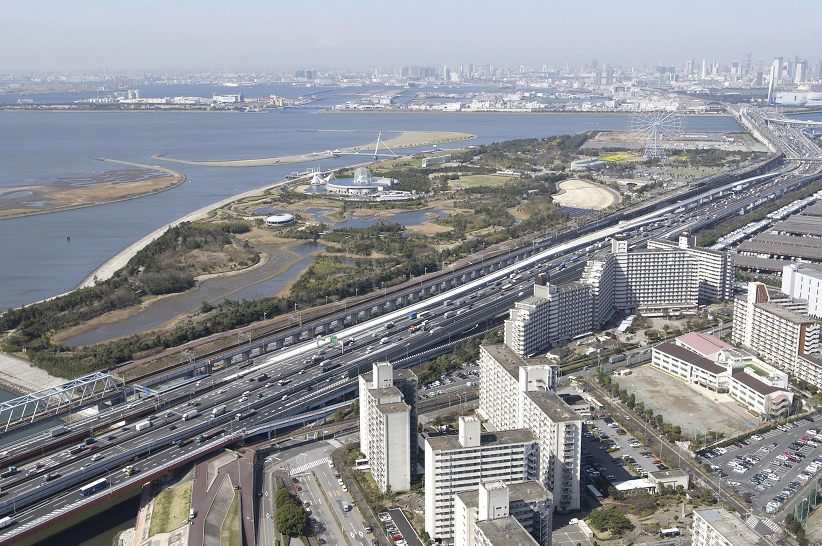 高速道路とマンション群の奥に広がる、海沿いの大きな公園と観覧車のある風景