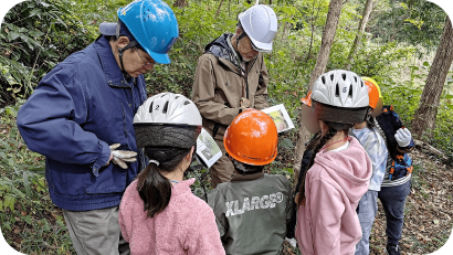 雑木林の中で、指導員とヘルメットを被った子供たちが円になって集まり、資料を見ながら話を聞いている様子。